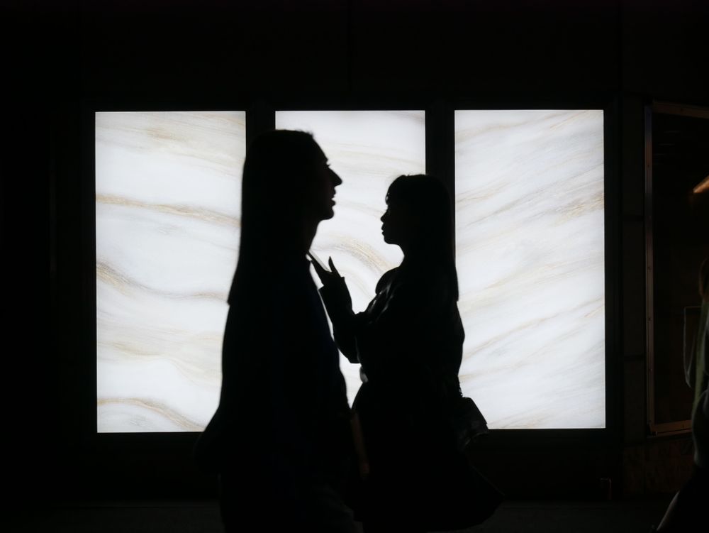 Two silhouetted figures standing against a brightly lit marble-patterned wall, creating dramatic contrast between dark figures and illuminated background